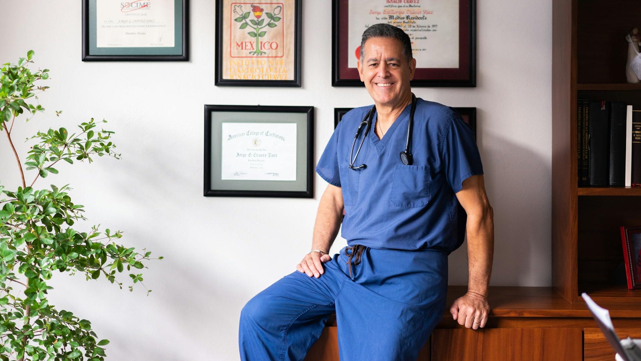 A smiling doctor in scrubs poses in his clinic, surrounded by framed certifications.
